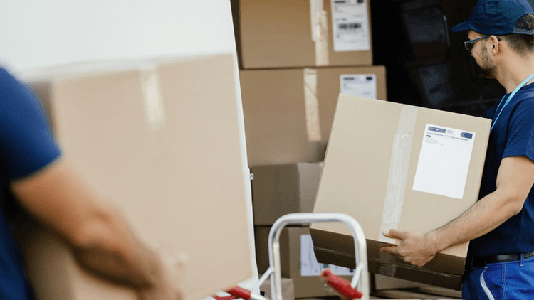 This image shows a worker unloading a large cardboard box from the back of a truck. The worker, wearing a blue shirt, glasses, and a cap, is carefully handling the box, likely as part of a logistics or delivery process. In the background, other boxes are stacked in the truck, ready to be unloaded. The visible shipping labels suggest that these are packages meant for delivery.