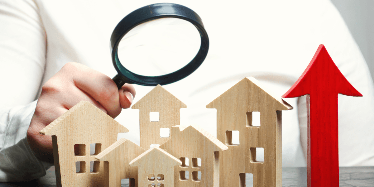 A hand holding a magnifying glass over wooden house figures and an upward arrow.