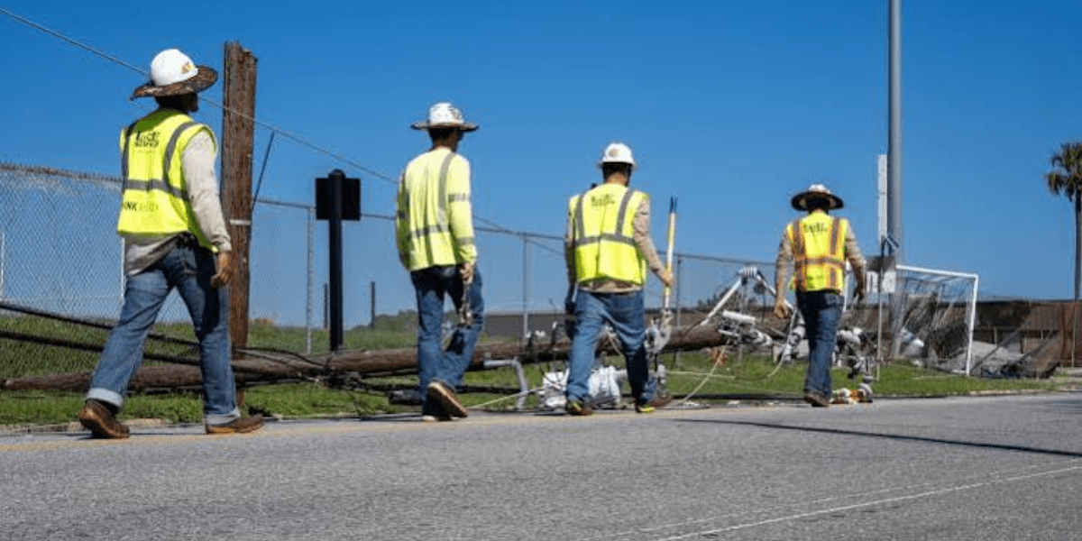 Four workers in safety vests walking past a fallen utility pole and debris
