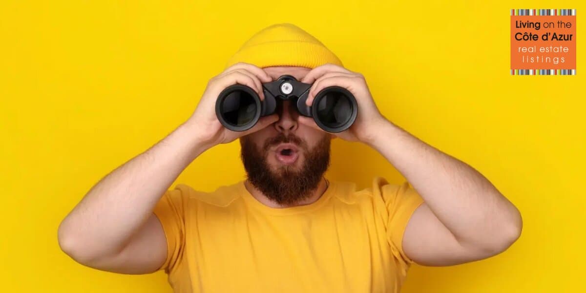 Man in yellow shirt and beanie looking through binoculars, symbolizing guided property search on the French Riviera.