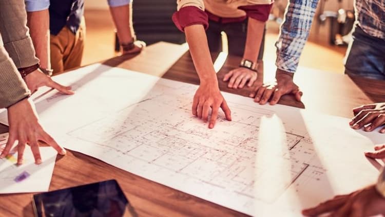 Multiple hands pointing at a blueprint during a collaborative meeting, likely related to a home remodeling project.