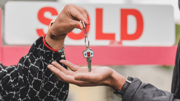 Person handing a house key to a buyer in front of a sold real estate sign.
