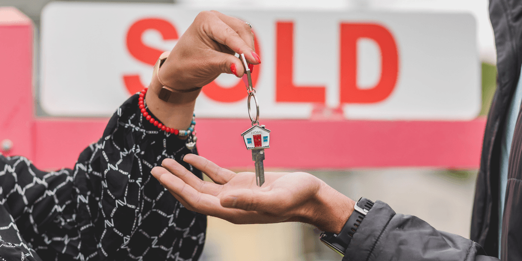 Person handing a house key to a buyer in front of a sold real estate sign.