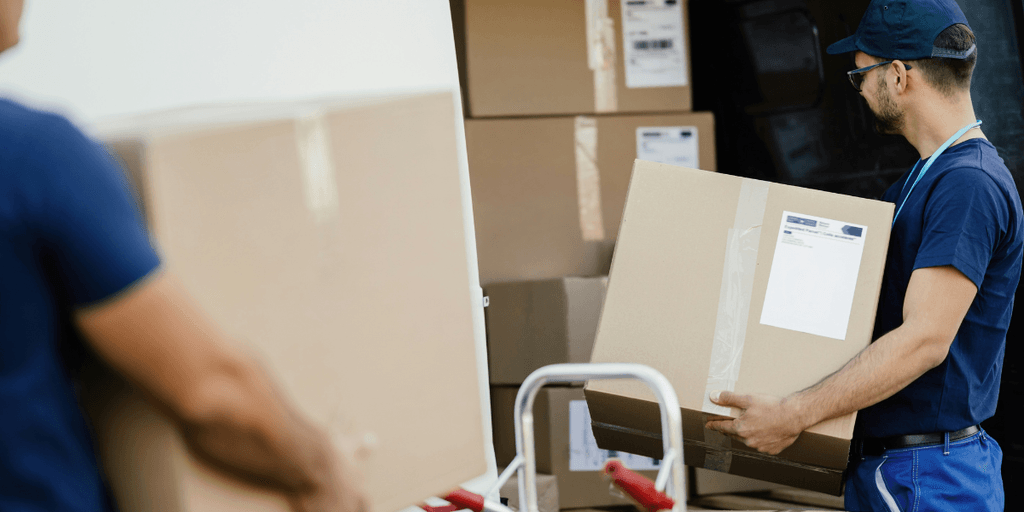 This image shows a worker unloading a large cardboard box from the back of a truck. The worker, wearing a blue shirt, glasses, and a cap, is carefully handling the box, likely as part of a logistics or delivery process. In the background, other boxes are stacked in the truck, ready to be unloaded. The visible shipping labels suggest that these are packages meant for delivery.