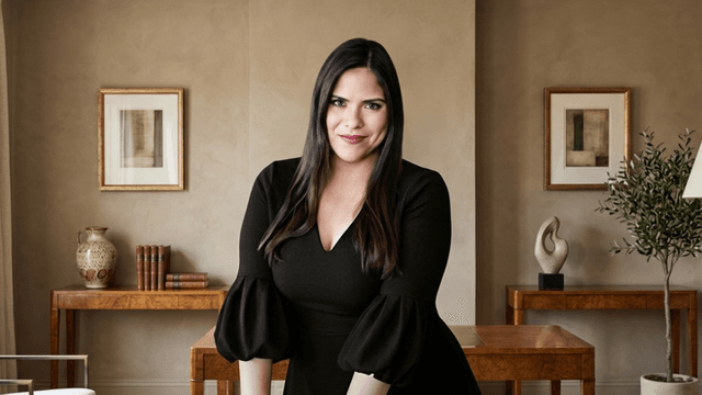 A woman with long dark hair stands confidently in a stylish, modern interior