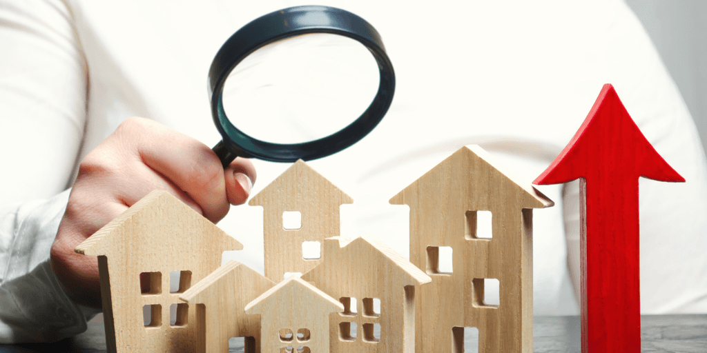 A hand holding a magnifying glass over wooden house figures and an upward arrow.
