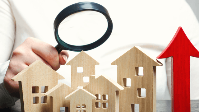 A hand holding a magnifying glass over wooden house figures and an upward arrow.