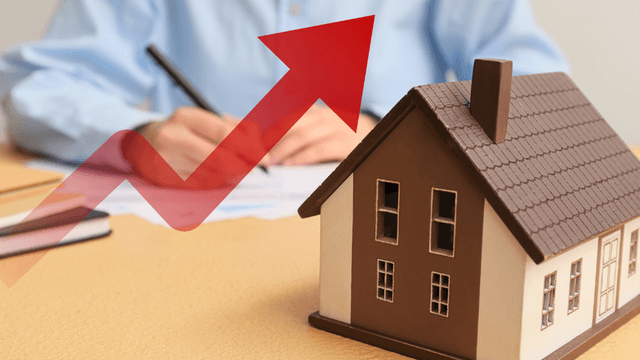 A miniature house model with an upward trend arrow and a person writing in the background