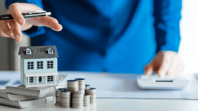 Person in a blue suit analyzing real estate investment with model house and coins
