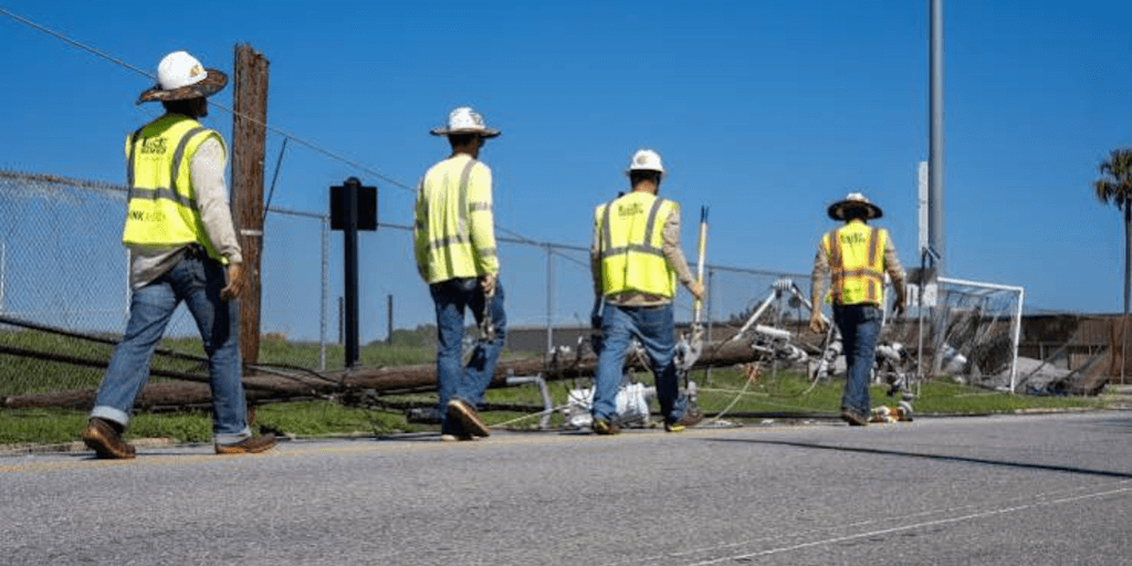 Four workers in safety vests walking past a fallen utility pole and debris