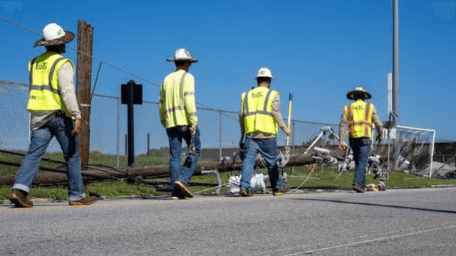 Four workers in safety vests walking past a fallen utility pole and debris