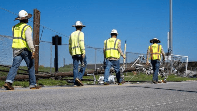 Four workers in safety vests walking past a fallen utility pole and debris