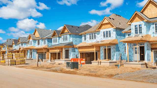 Newly constructed blue houses with wooden frames along a gravel road under a clear sky