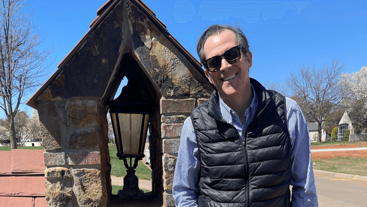 "Man wearing sunglasses standing next to a stone lamp post in a residential area on a sunny day, with trees and houses in the background."