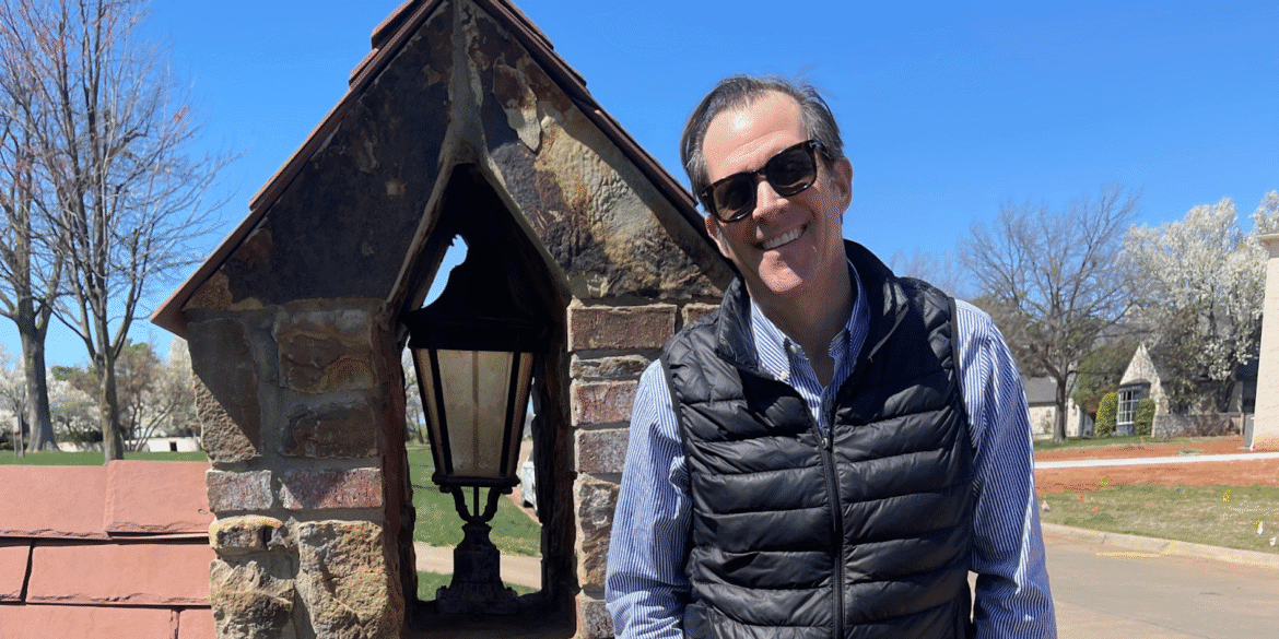 "Man wearing sunglasses standing next to a stone lamp post in a residential area on a sunny day, with trees and houses in the background."