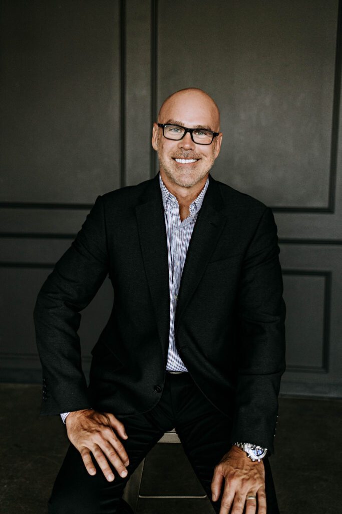 Wyatt Poindexter seated in a black suit against a dark paneled wall.